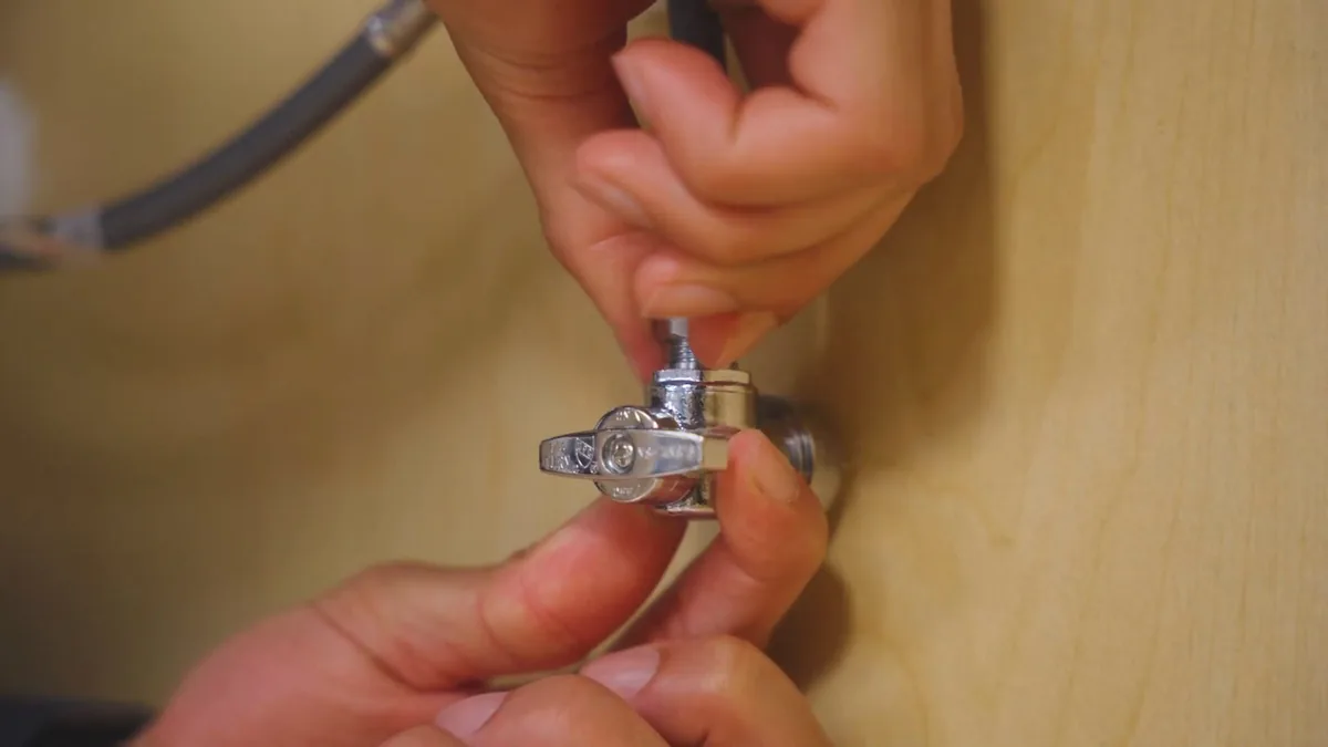 Hands holding a chrome angle stop valve on a wall inside a wood cabinet with a braided line