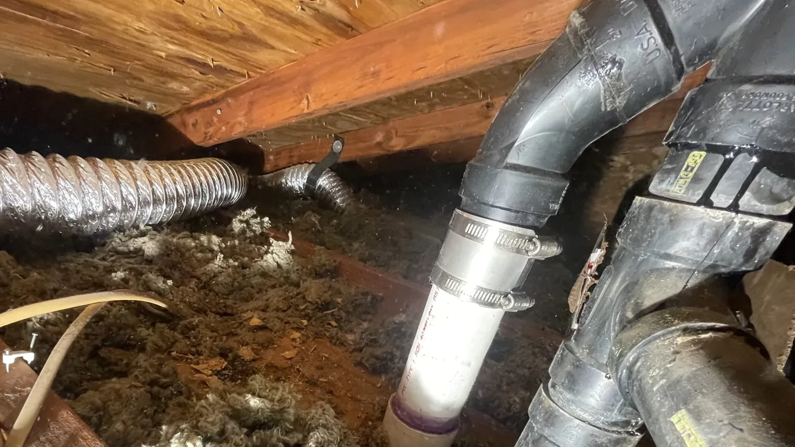 Attic space with gray drain pipes, a flexible duct and a bright pipe fitting among insulation