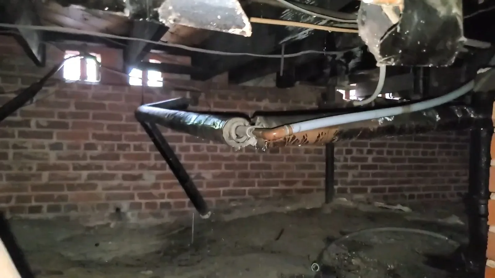 Crawlspace with a brick wall, small windows and a black drain pipe running across old joists