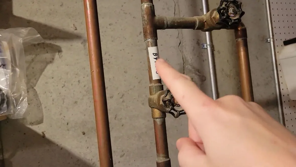 Finger pointing at a valve on copper supply lines along a basement wall