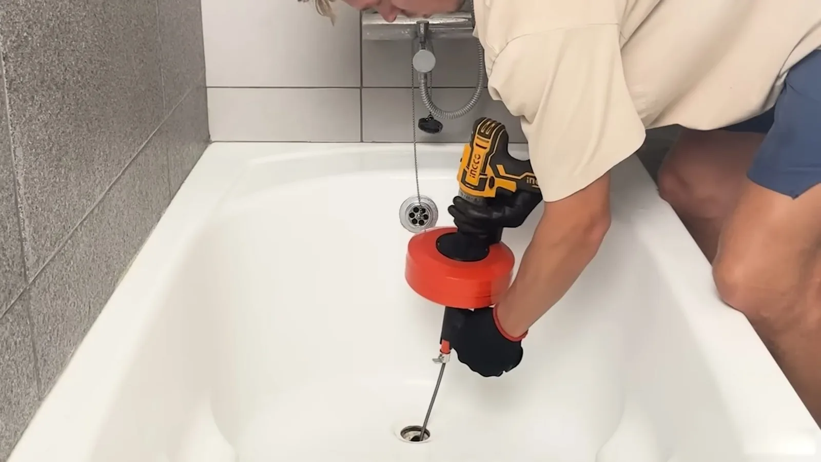 Person in a tan shirt feeding a drill-driven auger into a white bathtub drain