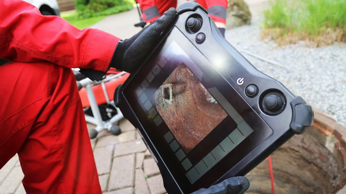 Worker in red coveralls holding a handheld monitor that shows a pipe camera view