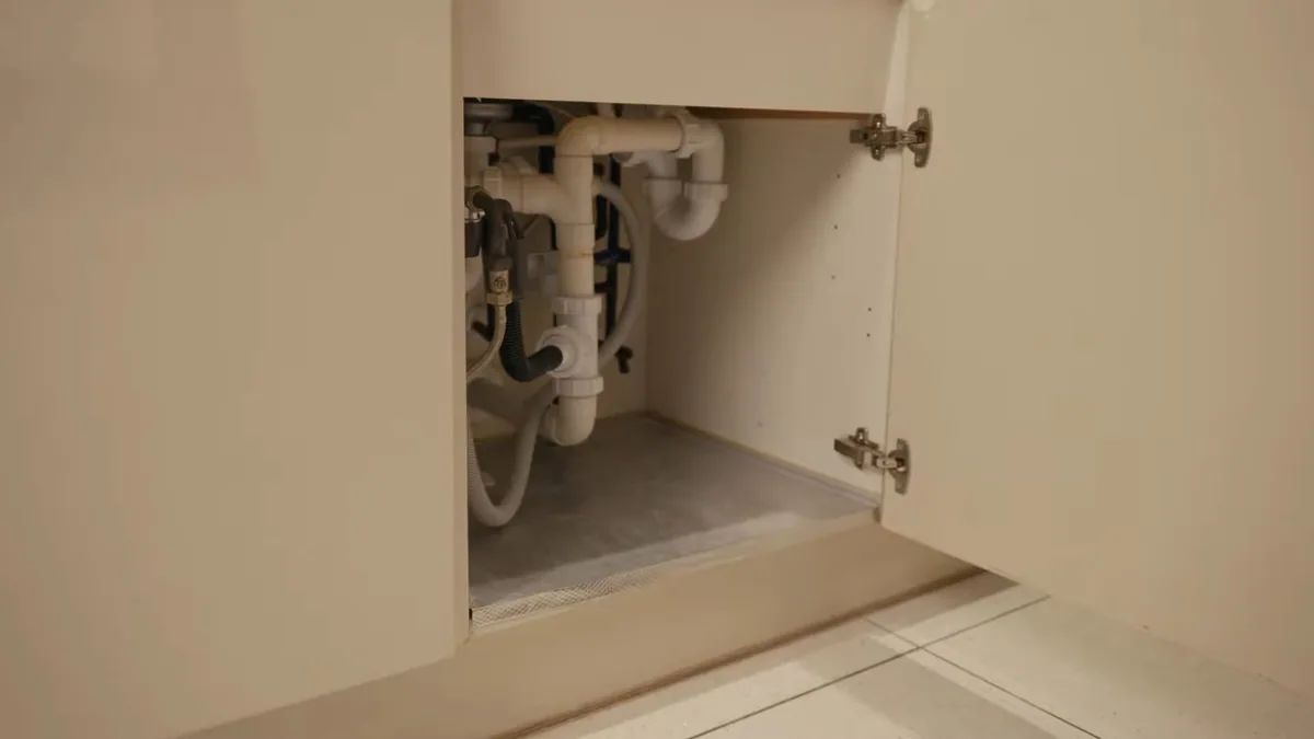 Open sink cabinet with white drain pipes and a trap behind the cabinet door