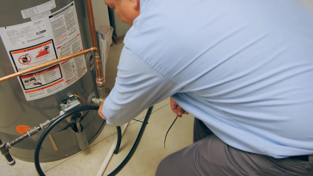 Man in a light blue shirt crouching at a tank water heater with a black drain hose attached