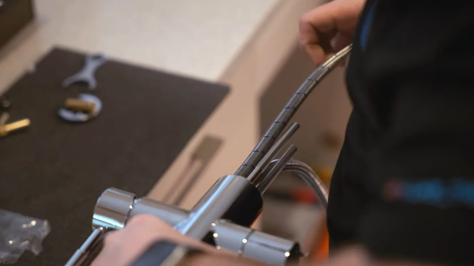 Chrome faucet body with a braided hose attached and a hand beneath it on a dark counter, wrenches in the background