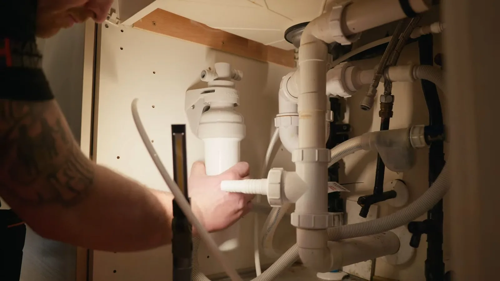 Hand reaching into a cabinet toward white drain fittings and pipes under a kitchen sink