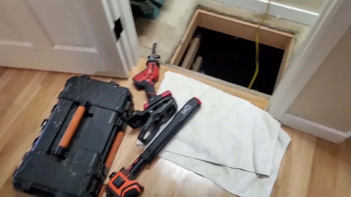 Open tool case, pipe wrench and reciprocating saw on a towel beside a floor access hatch