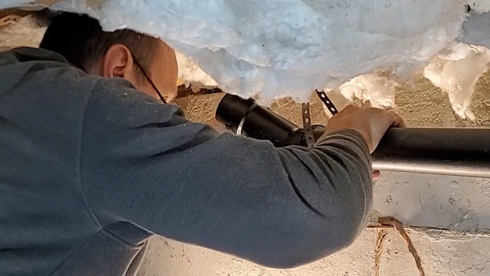 Technician reaching up to a dark drain line in a crawl space under hanging insulation