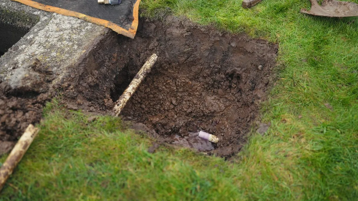 Square hole dug in a grassy lawn beside a concrete slab, with a wooden stake lying across the soil and a short pipe stub at the bottom
