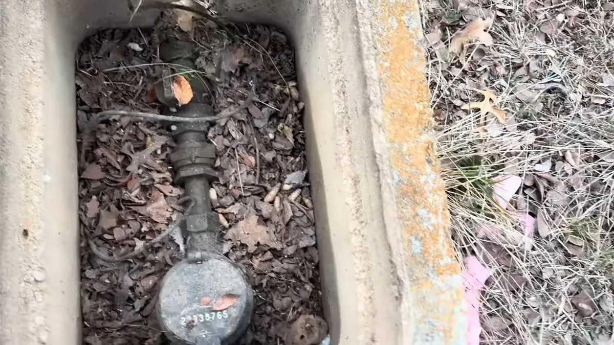 Water meter with a numbered register at the bottom of a concrete box, covered in dry leaves