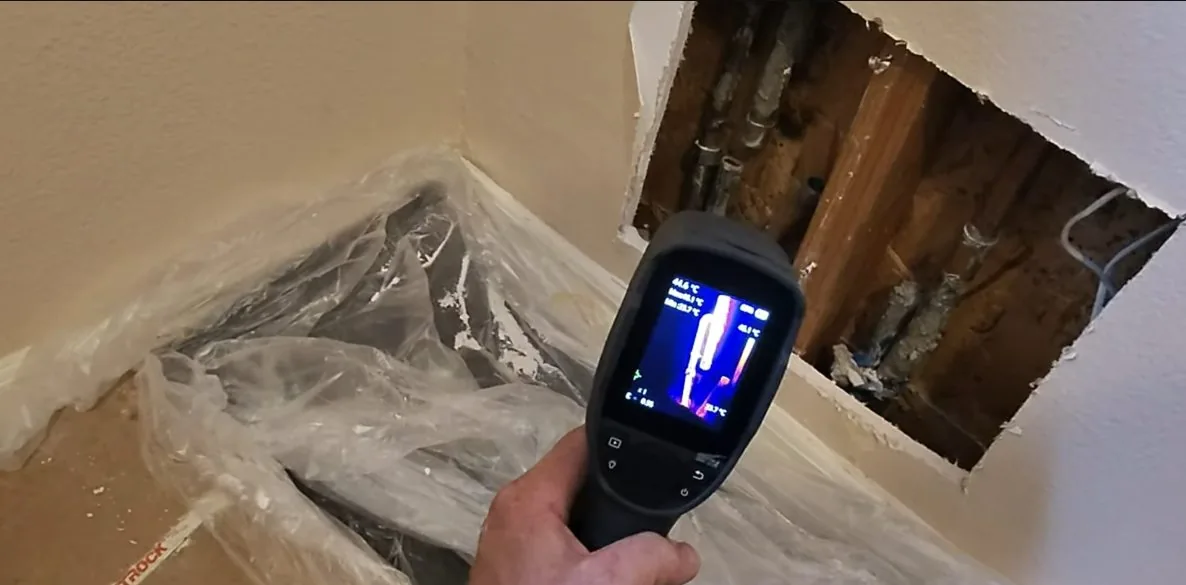 Hand holding a thermal camera toward an opened wall cavity, its screen showing a bright vertical heat pattern