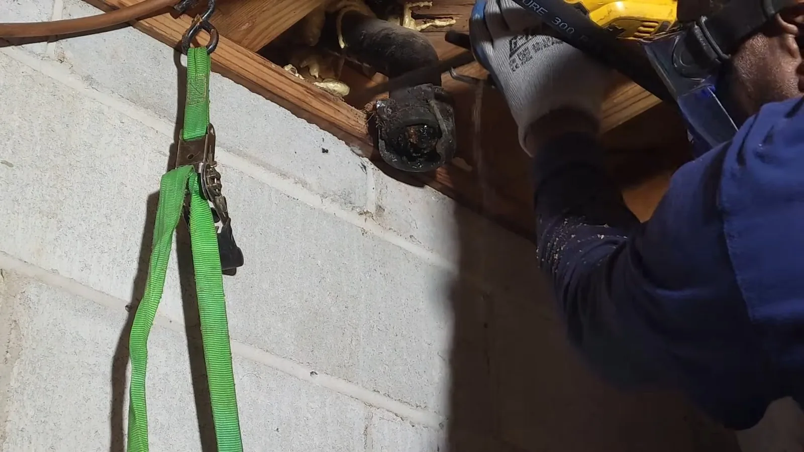 Man in a blue jacket and work gloves cutting a pipe at a floor joist above a block wall, a green strap hanging beside him