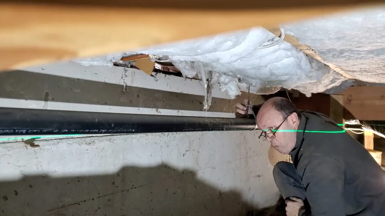 Man with glasses kneeling under a floor beam with a green laser line and insulation hanging above