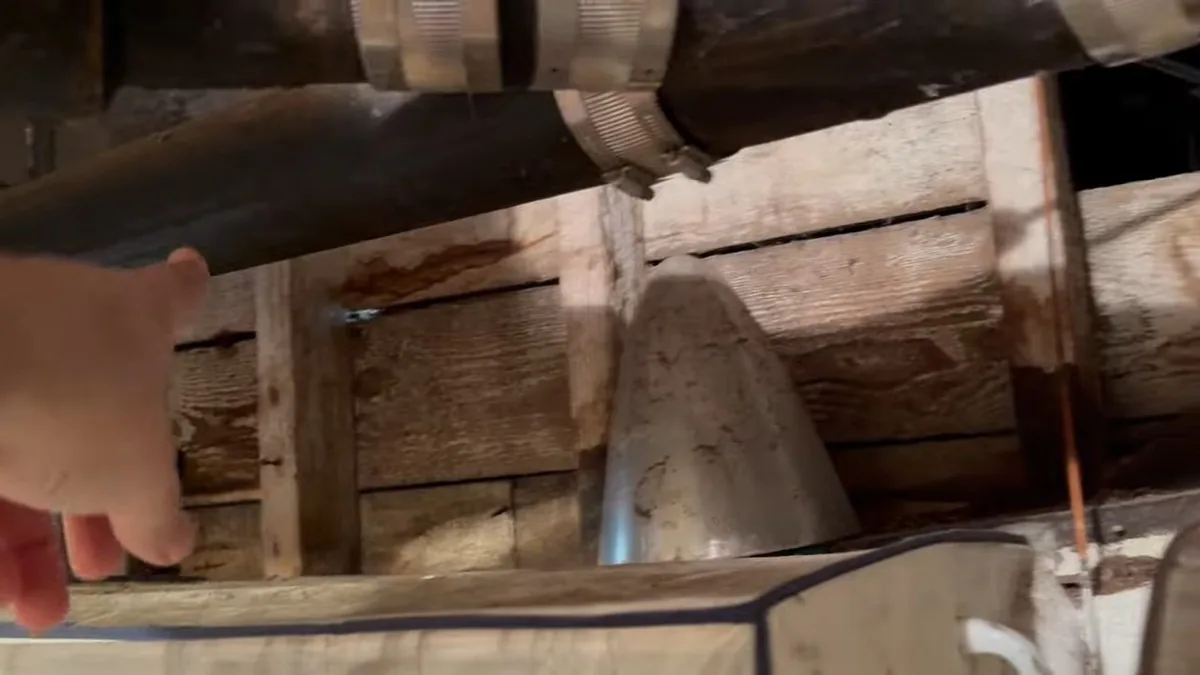 Old cast iron drain pipe running under a wood floor frame with a hand reaching in