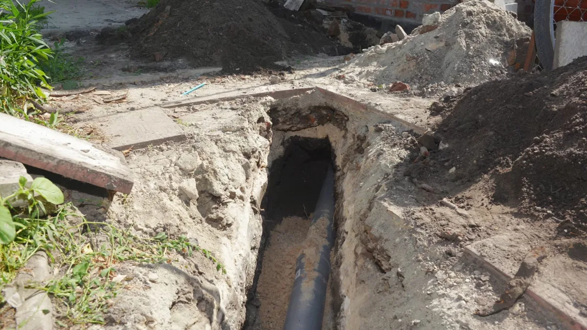 Narrow trench dug through sandy soil beside a house with a dark pipe exposed along the bottom