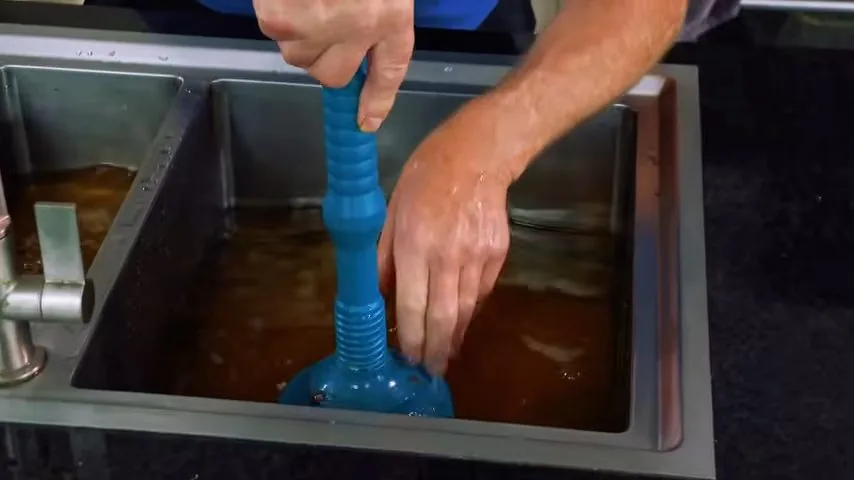 Two hands working a blue plunger in a stainless steel sink filled with dirty brown water