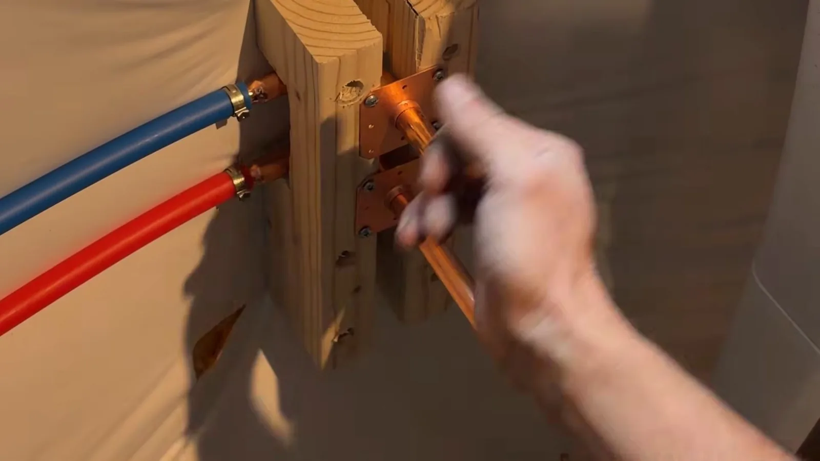 Red and blue PEX lines clamped to a wooden stud in an open wall with a hand working a copper crimp fitting