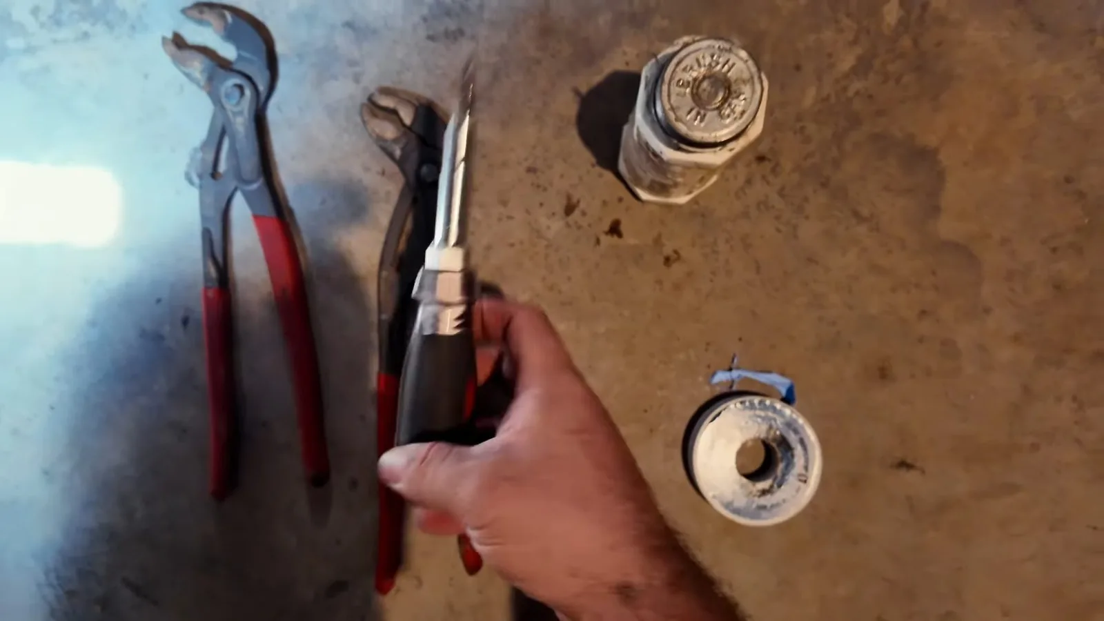 Slip-joint pliers, a screwdriver and a spool of thread tape laid on dirt beside a pipe stub, with a hand holding a tool