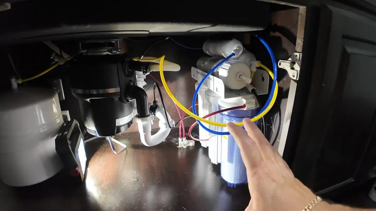 Cabinet under a sink holding a disposal, a white drain trap and a filter system with colored tubing