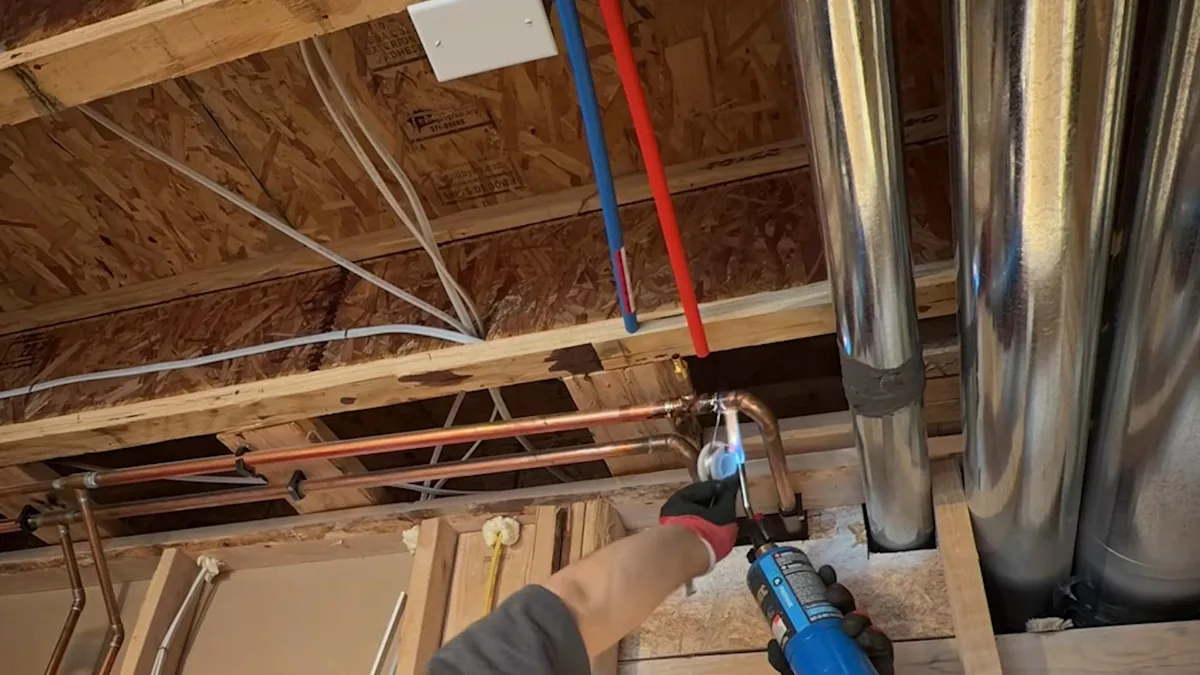 Red and blue PEX and copper pipe running along ceiling joists while a hand holds a torch