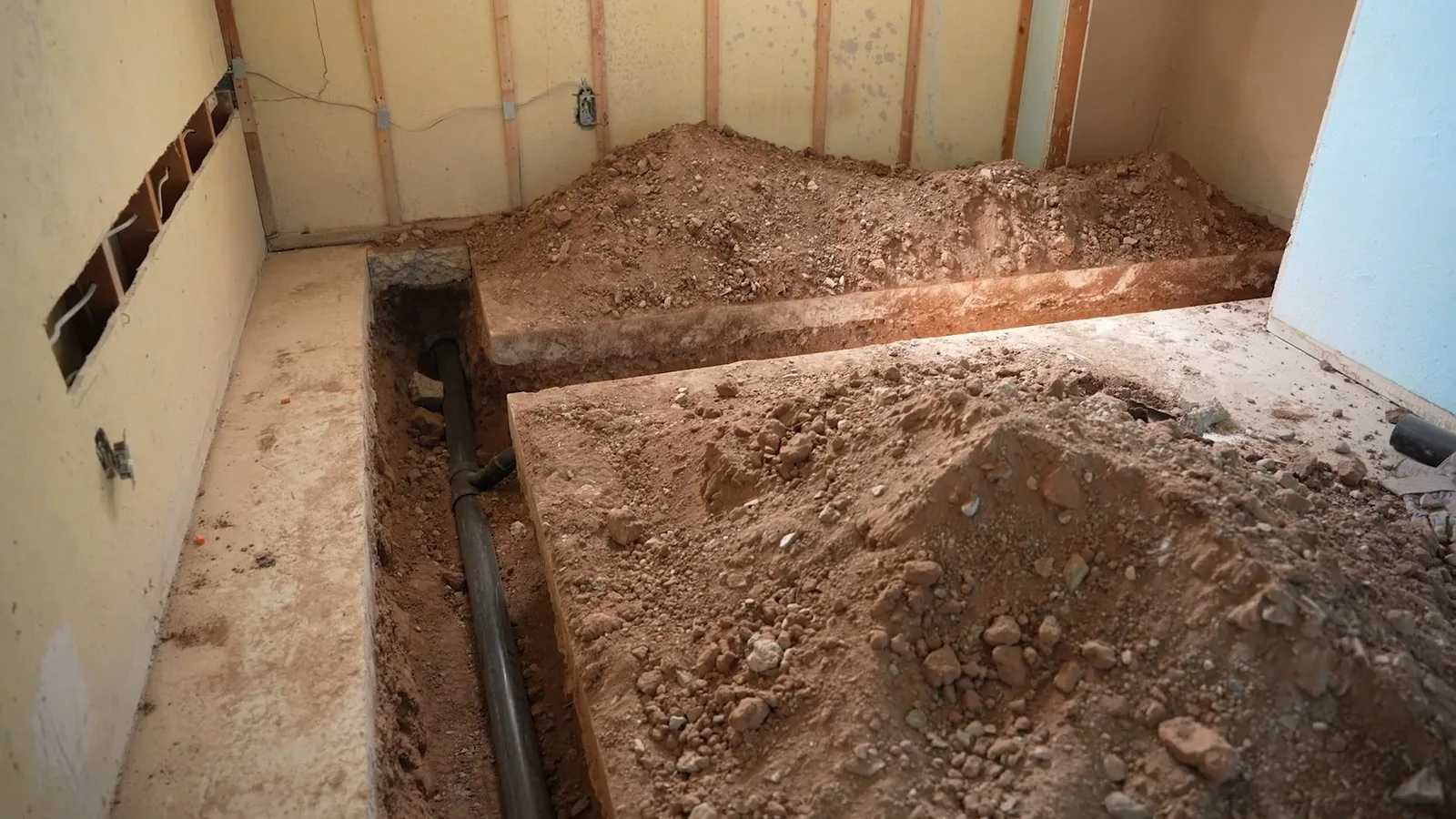 Trench cut through a house floor with a dark pipe lying in the soil and piles of dirt along the wall