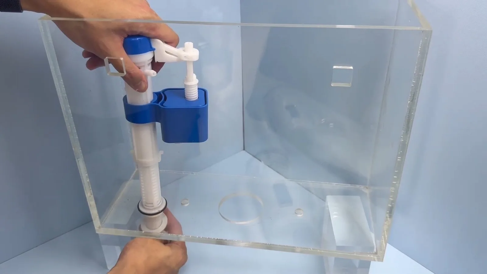 Hands adjusting a blue fill valve inside a clear acrylic toilet tank