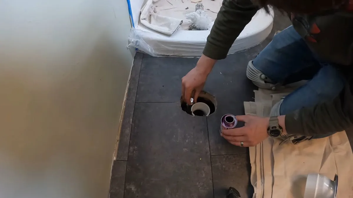 Person crouched over a round drain opening in a bathroom subfloor with a tube in one hand