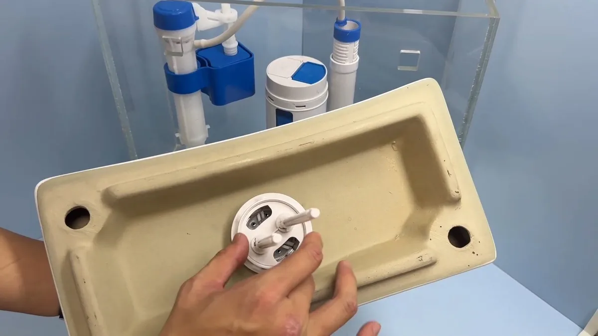 Hands holding a toilet tank lid upside down beside a clear tank with a blue fill valve