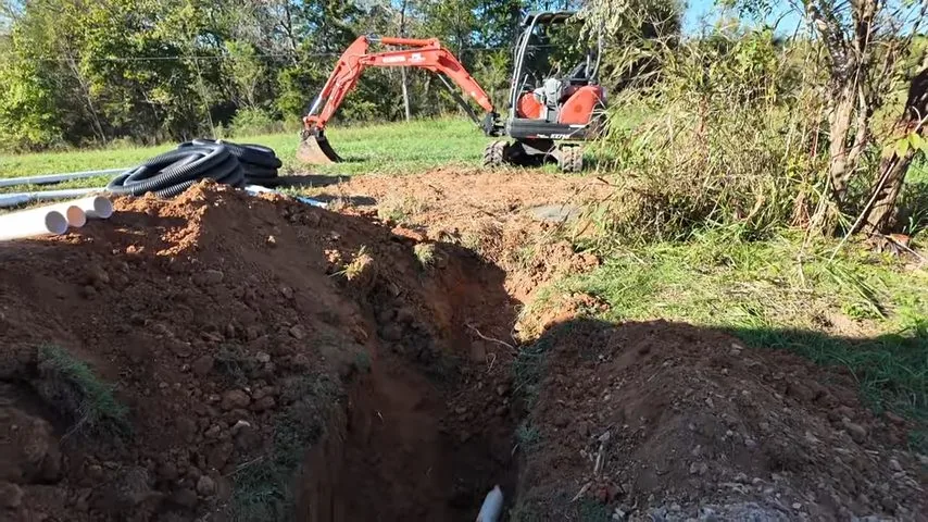 Small orange excavator behind a long open trench with black corrugated and white pipe lying beside it