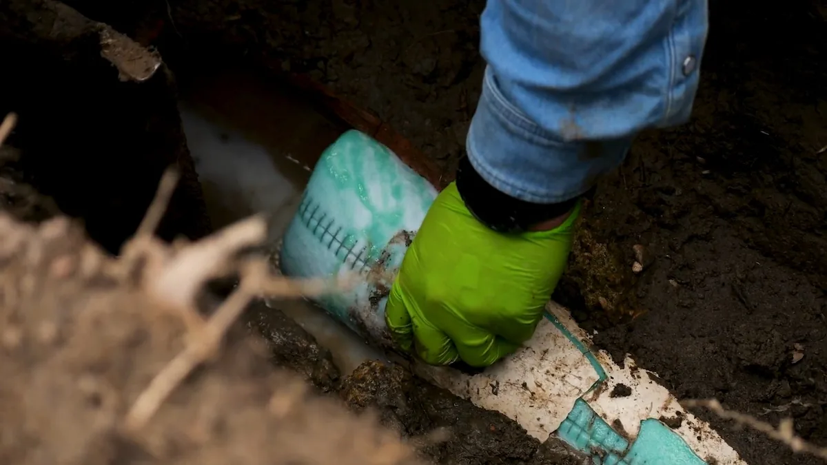 Gloved hand pressing a white and teal liner sleeve against a pipe in a dirt pit