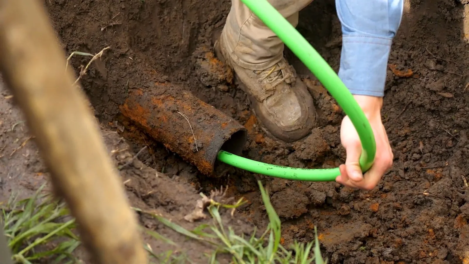 Hand feeding a green cable into a rusted pipe end exposed in a dug trench