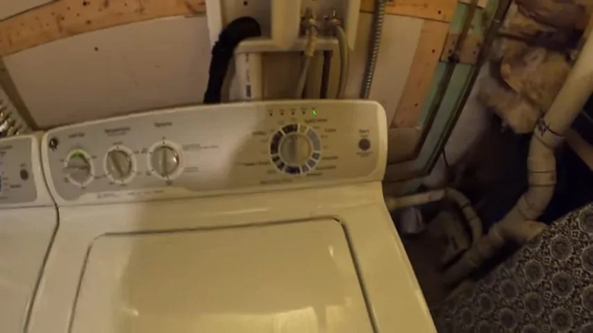 Cream top-load washer with hoses and pipes in the wall behind it