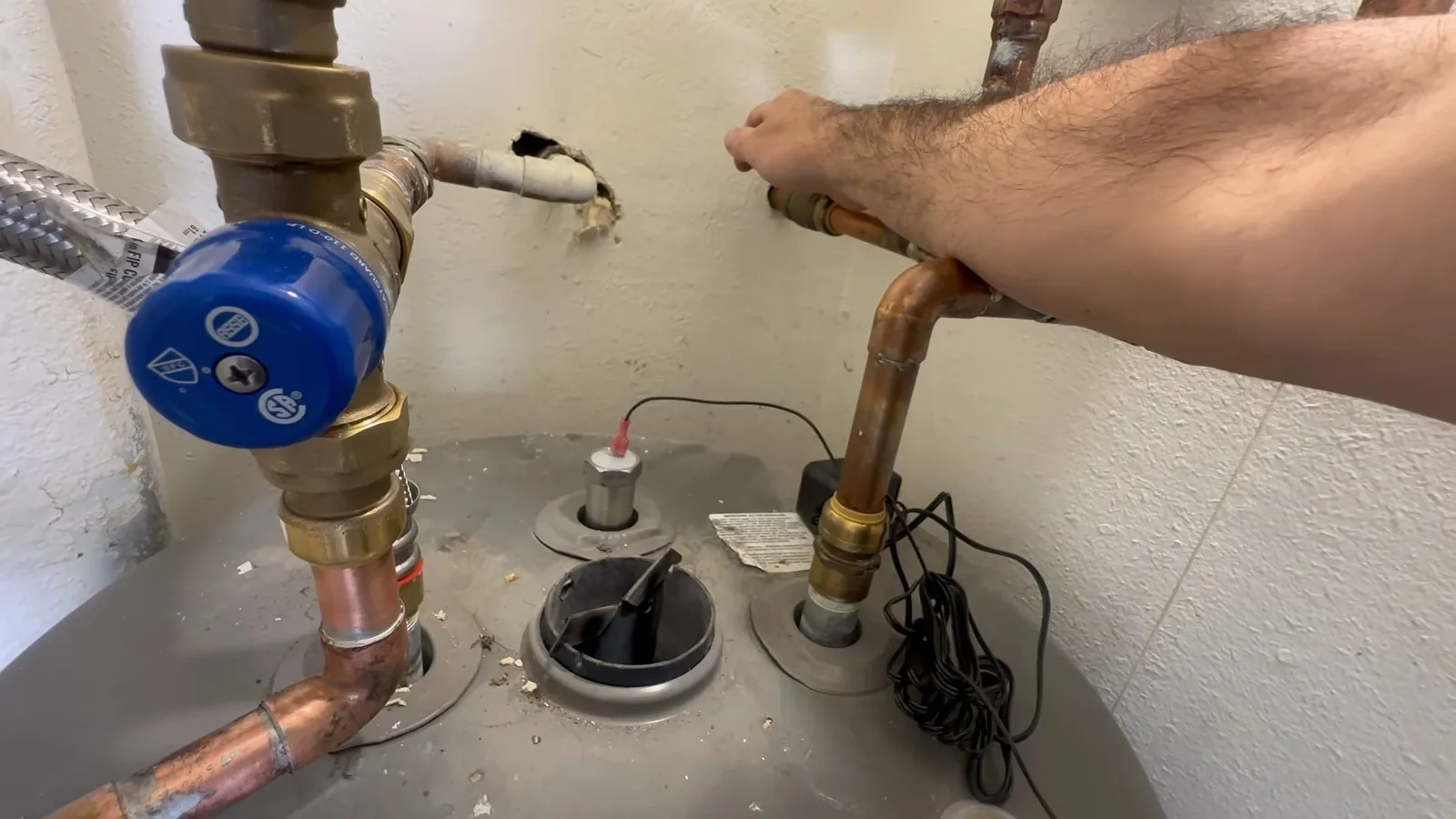Hand reaching across the top of a tank water heater with copper lines, a flexible connector and a blue valve