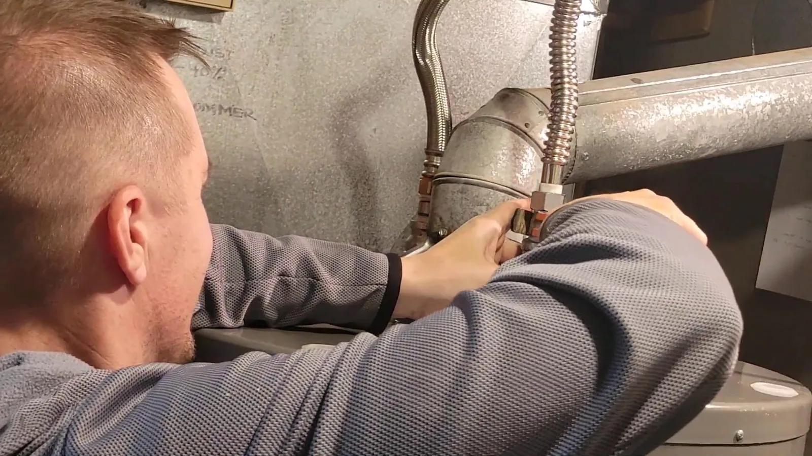 Man in a gray sweater working at the metal flue and gas line on top of a water heater