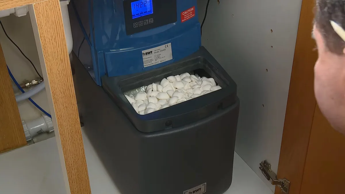 Man looking into an open softener brine tank holding white salt tablets under a cabinet