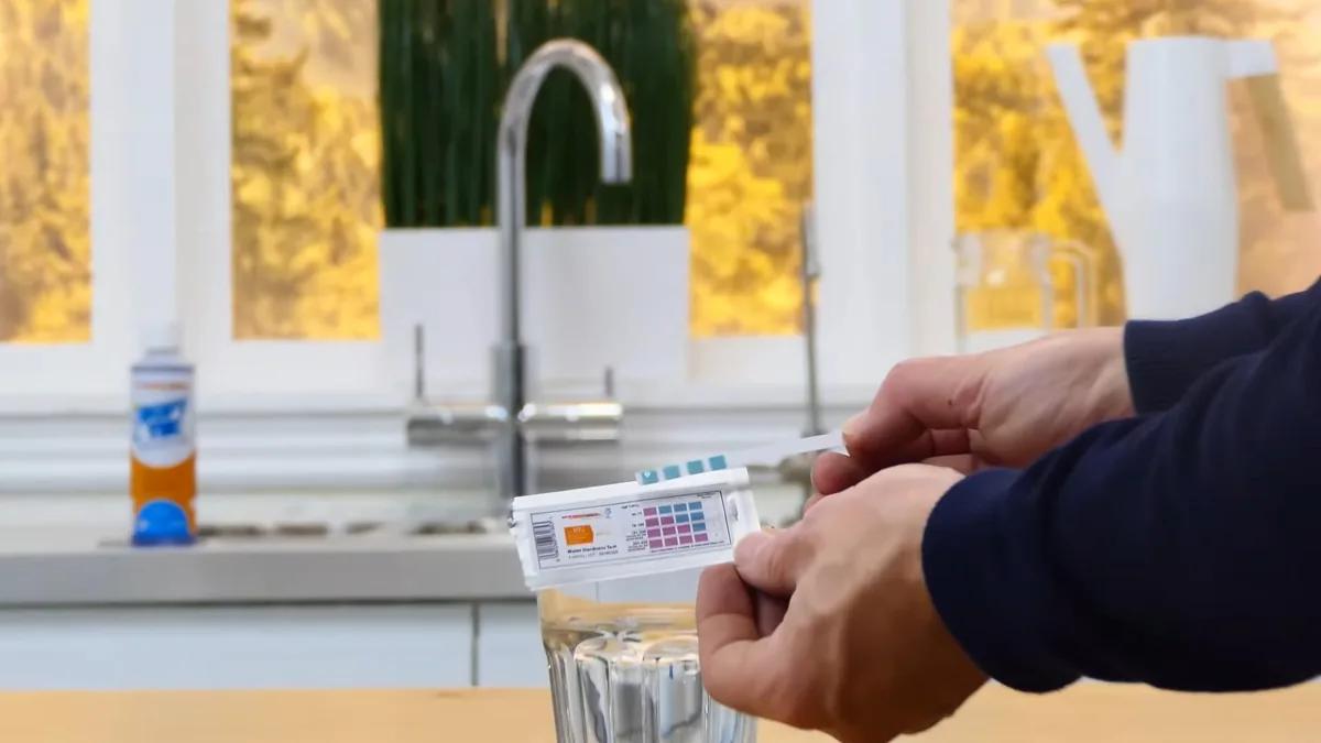 Hands holding a color test strip over a glass of water at a kitchen faucet
