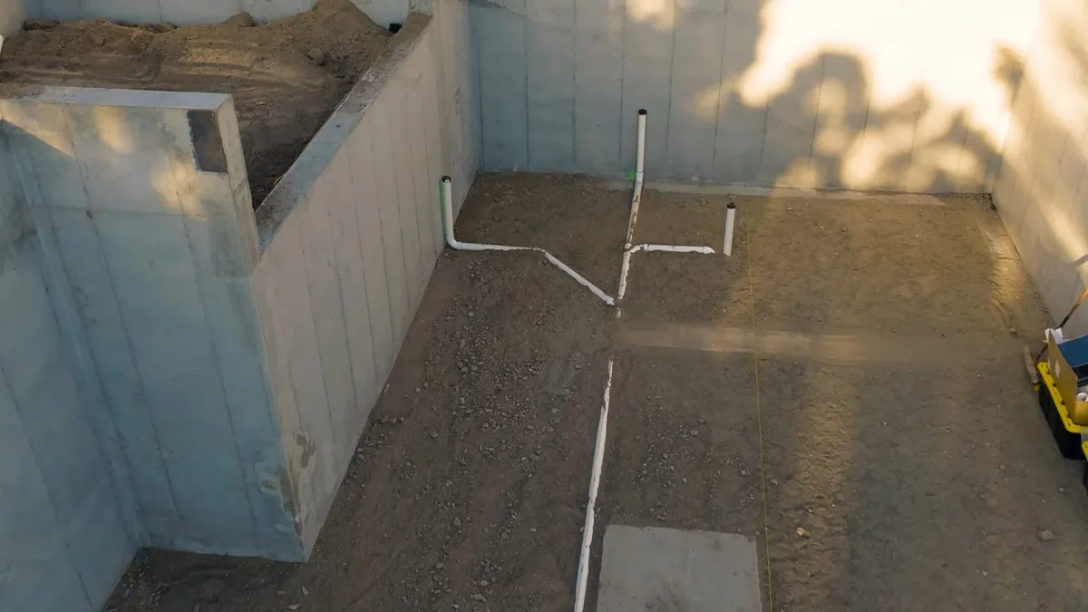 Dirt-floor area inside low block walls with white drain pipes laid out in a pattern
